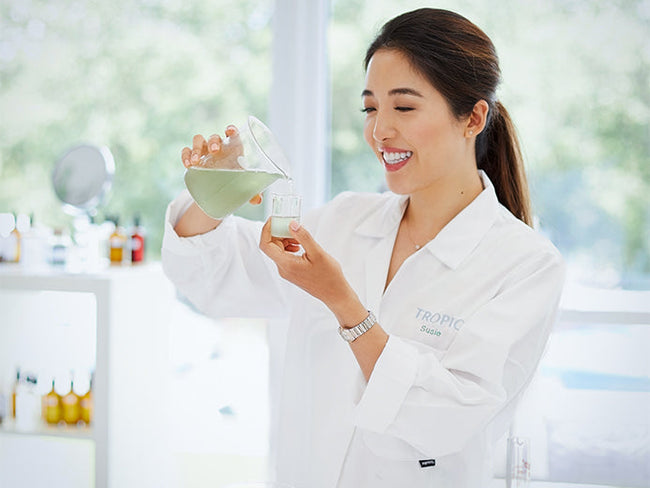 Image shows Susie in a lab coat pouring light green liquid from a beaker into a smaller glass beaker. There is a table in the background with products on and windows looking out to green trees.